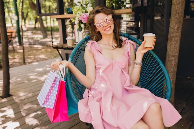 Free photo portrait of young smiling happy attractive woman sitting in cafe with shopping bags drinking coffee, summer fashion outfit, pink cotton dress, trendy apparel