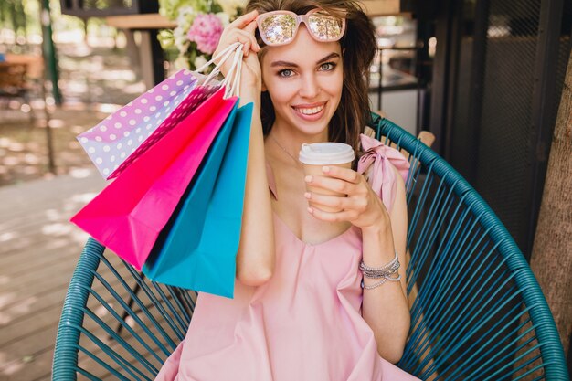 Ritratto di giovane donna graziosa felice sorridente con espressione faccia eccitata seduto nella caffetteria con borse della spesa bere caffè, vestito di moda estiva, abito di cotone rosa, abbigliamento alla moda