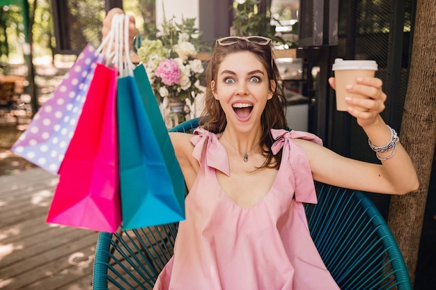 Foto gratuita ritratto di giovane donna graziosa felice sorridente con espressione faccia eccitata seduto nella caffetteria con borse della spesa bere caffè, vestito di moda estiva, abito di cotone rosa, abbigliamento alla moda