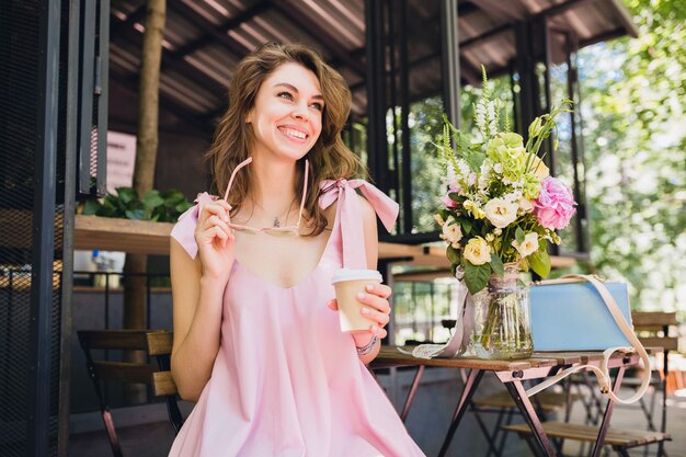 Portrait of young smiling happy pretty woman with sitting in cafe drinking coffee, summer fashion outfit, pink cotton dress, trendy apparel accessories