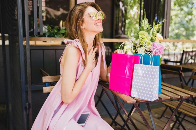 Portrait of young smiling happy pretty woman with surprised face expression sitting in cafe with shopping bags, summer fashion outfit, pink cotton dress, trendy apparel