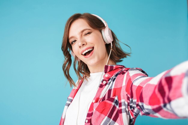 헤드폰으로 음악을 듣고 분홍색 배경 위에 카메라를 행복하게 보고 있는 셔츠를 입은 웃고 있는 젊은 여성의 초상화