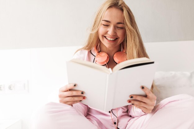 Portrait of young smiling lady sitting on bed with book in hands and laughing at home