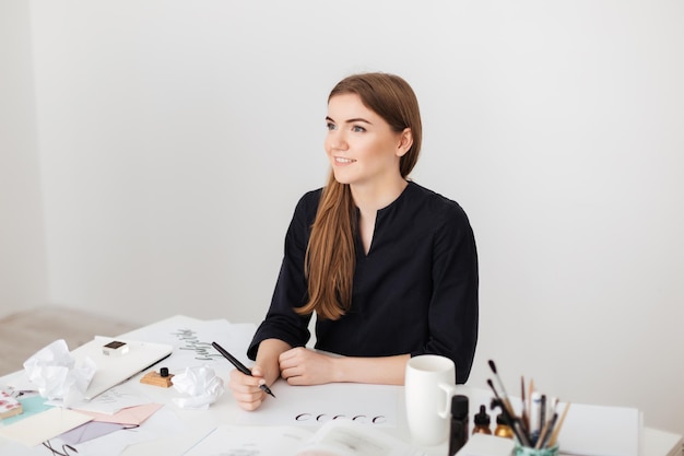 Portrait of young smiling lady sitting at the white desk and writing notes on paper while dreamily looking aside isolated