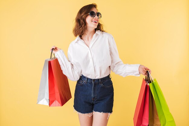 Portrait of young smiling lady in sunglasses standing with colorful shopping bags in hands on over pink background. Pretty woman standing in white shirt and denim shorts