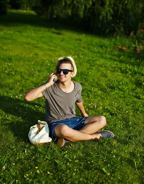 portrait of young smiling laughing attractive modern stylish man in casual cloth in hat in glasses sitting in the park in green grass speaking on phone