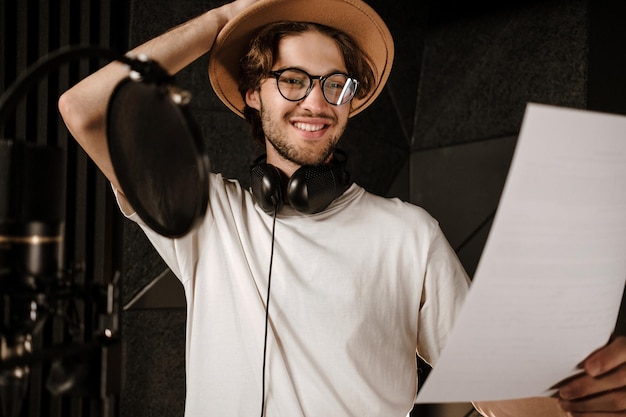Portrait of young smiling male singer happily reading text of new song in modern recording studio