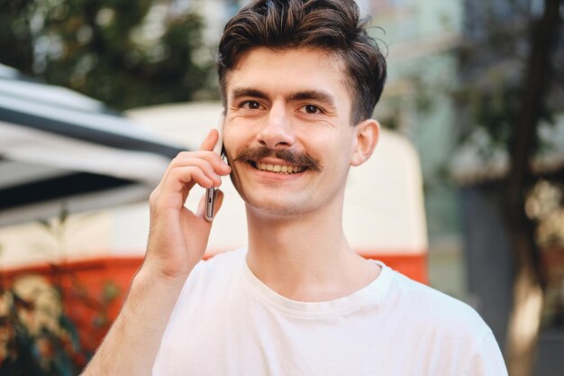 Portrait of young smiling mustache man in withe T-shirt talking on cellphone while happily looking in camera on city street