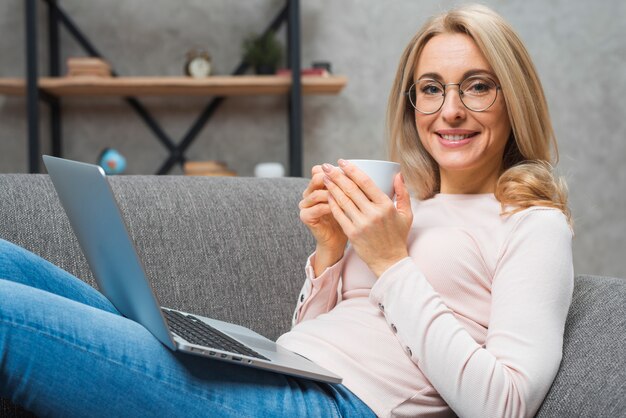 Портрет молодой улыбающейся женщины, держащей чашку кофе с открытым ноутбуком на коленях