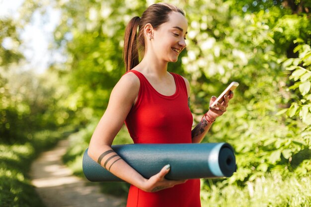 Portrait of young smiling woman in red jumpsuit standing with yoga mat in hand and using cellphone in park