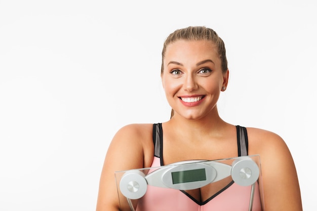 Free photo portrait of young smiling woman with excess weight holding scales in hands joyfully looking in camera over white background isolated