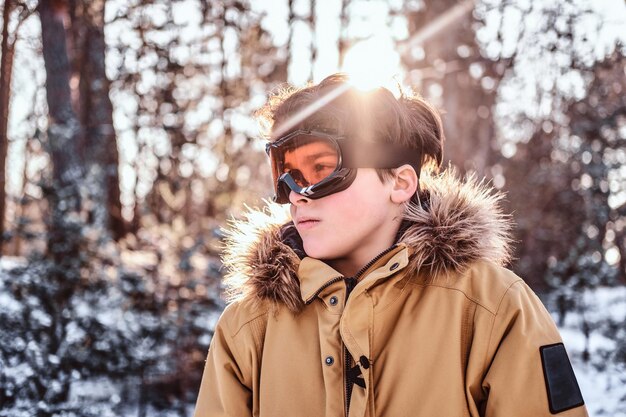 일출 동안 눈 덮인 숲에 서 있는 따뜻한 옷과 보호용 고글을 입은 젊은 스노보더의 초상화