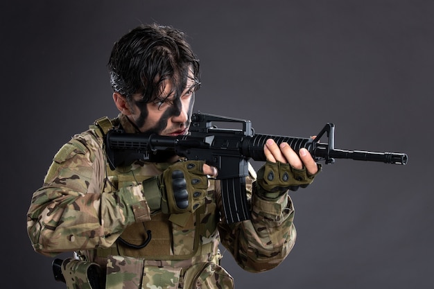 Portrait of young soldier in camouflage aiming machine gun on dark wall