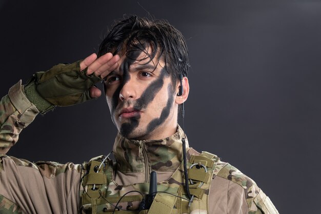 Portrait of young soldier in camouflage saluting on a dark wall