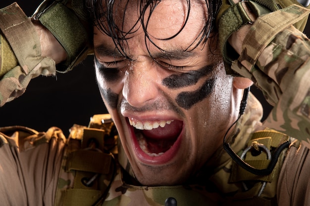 Portrait of young soldier in camouflage screaming from pain on black wall