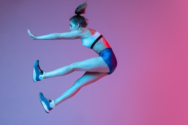 Portrait of young sportive girl in sportswear training jumping isolated over pink studio background in neon light