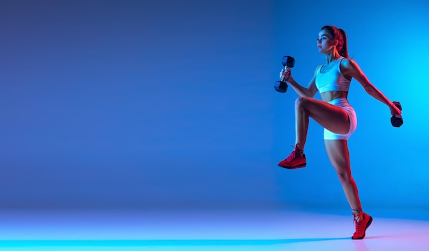 Портрет молодой спортивной девушки, тренирующейся с гантелями на синем фоне в неоне