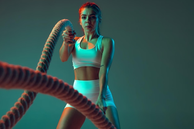 Portrait of young spotive girl doing exercises with rope keeping body fit isolated over green background in neon