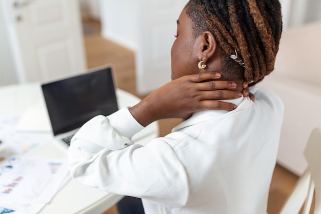 PCで作業した後、肩の痛みに苦しんでいる痛みを伴う表情で痛む肩に触れるラップトップの前にホームオフィスの机に座っている若いストレスの多いアフリカの女性の肖像画