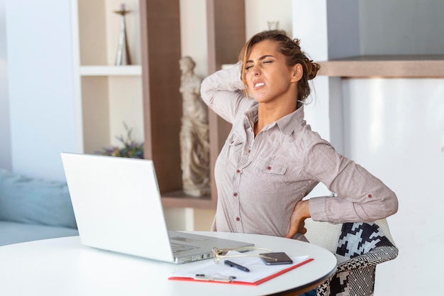 Portrait of young stressed woman sitting at home office desk in front of laptop touching aching back with pained expression suffering from backache after working on laptop