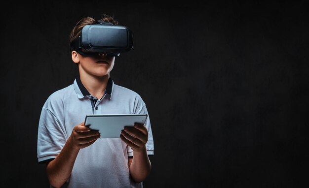 가상 현실 안경과 태블릿 컴퓨터를 사용하여 흰색 티셔츠를 입은 어린 학생 소년의 초상화. 어두운 배경에 고립.