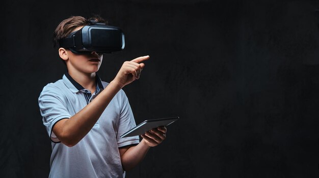 Portrait of a young student boy dressed in a white t-shirt using virtual reality glasses and tablet computer. Isolated on a dark background.