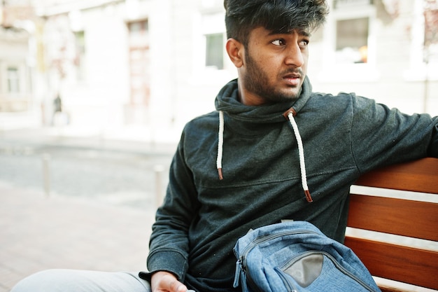 Портрет молодого стильного индийского мужчины-модели позирует на улице, сидя на скамейке и держа смартфон под рукой