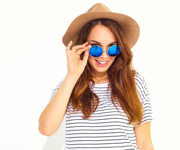 白い壁に分離された茶色の帽子でカジュアルな夏服の若いスタイリッシュな笑う女性モデルの肖像画