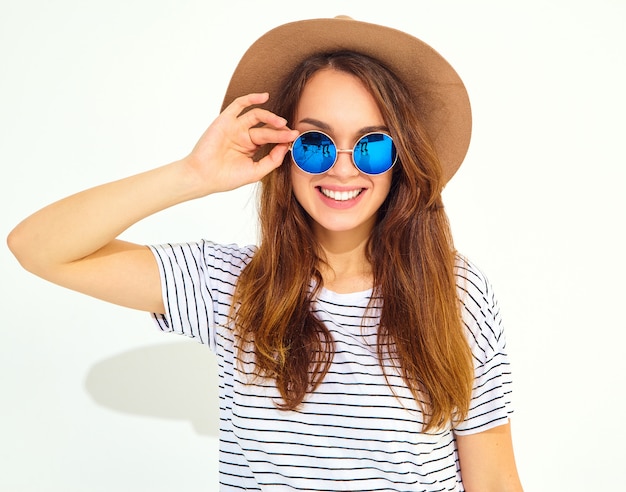 白い壁に分離された茶色の帽子でカジュアルな夏服の若いスタイリッシュな笑う女性モデルの肖像画
