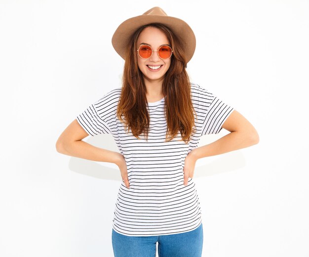 白い壁に分離された自然なメイクと茶色の帽子でカジュアルな夏服の若いスタイリッシュな笑う女性モデルの肖像画。