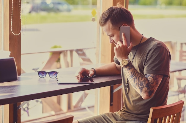 Portrait of young stylish man with tattooed arms talks by smartphone indoor.