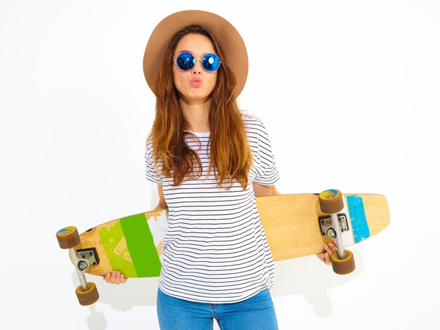 Portrait of young stylish woman model in casual summer clothes in brown hat posing with longboard desk. Isolated on white
