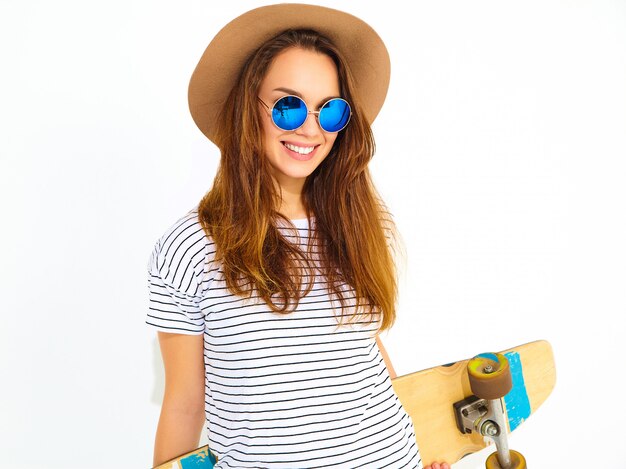 Portrait of young stylish woman model in casual summer clothes in brown hat posing with longboard desk. Isolated on white