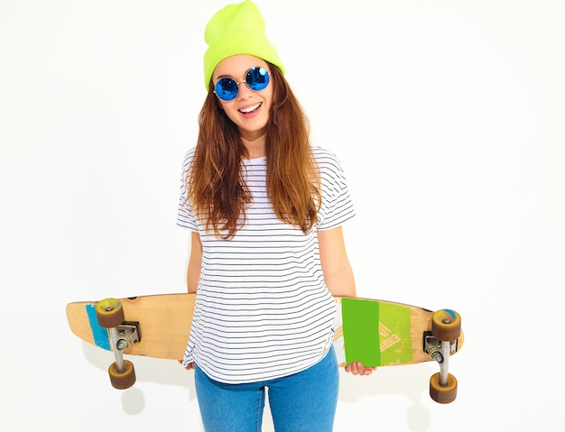 Portrait of young stylish woman model in casual summer clothes in yellow beanie hat posing with longboard desk. Isolated on white