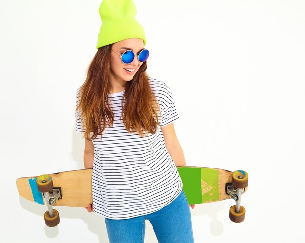 Portrait of young stylish woman model in casual summer clothes in yellow beanie hat posing with longboard desk. Isolated on white