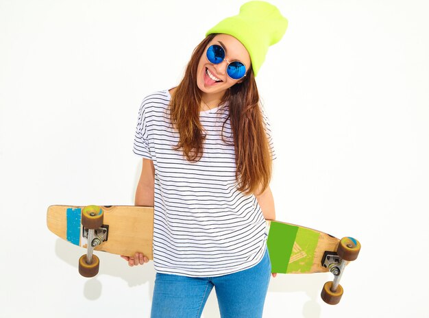 Portrait of young stylish woman model in casual summer clothes in yellow beanie hat posing with longboard desk. Isolated on white