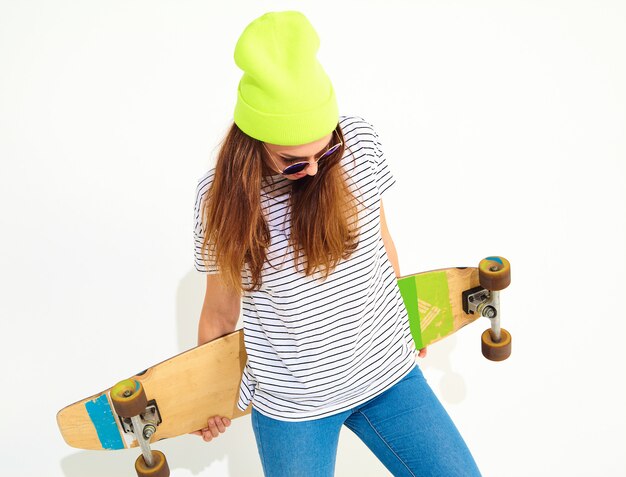 Portrait of young stylish woman model in casual summer clothes in yellow beanie hat posing with longboard desk. Isolated on white