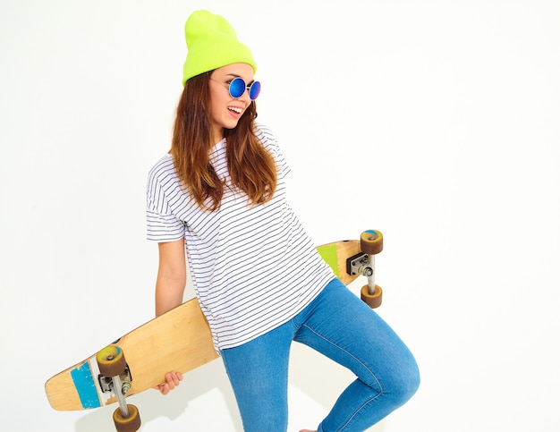 Portrait of young stylish woman model in casual summer clothes in yellow beanie hat posing with longboard desk. Isolated on white