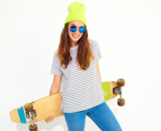 Portrait of young stylish woman model in casual summer clothes in yellow beanie hat posing with longboard desk. Isolated on white