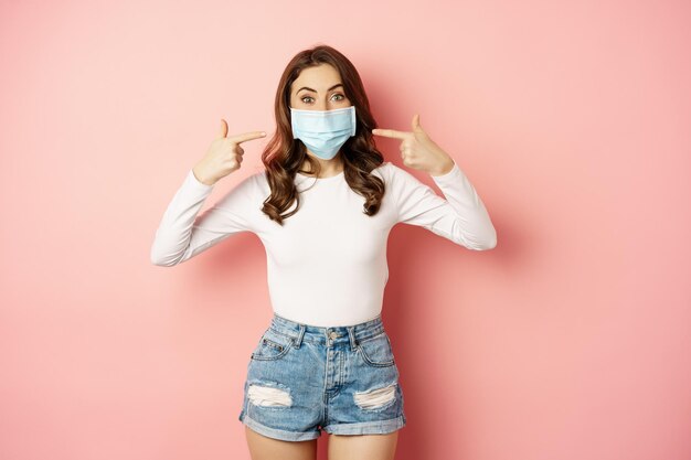 Portrait of young stylish woman wearing and pointing fingers at her medical face mask smiling preven...