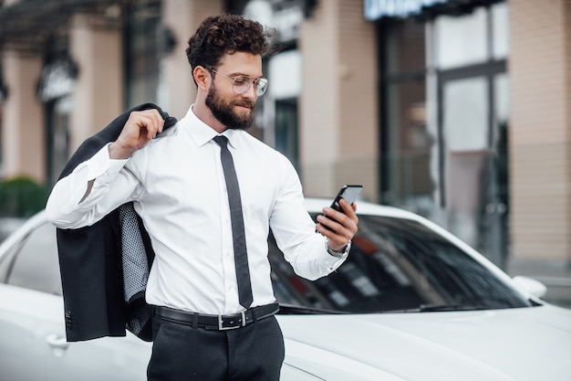 Portrait of a young, successful, handsome man in a suit on the background of a new white car, reading mail on his smartphone