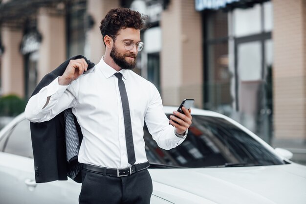 彼のスマートフォンでメールを読んで、新しい白い車の背景にスーツを着た若い、成功した、ハンサムな男の肖像画