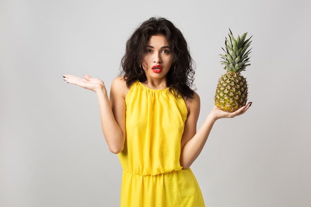 Portrait of young surprised pretty woman in yellow dress, holding pineapple, funny emotion, shocked face expression, summer style, fruit diet, looking in camera, mixed race, isolated, holding hands up