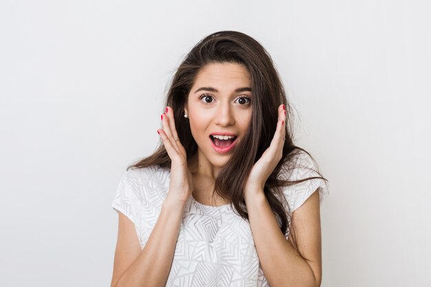 Portrait of young surprised woman in white blouse with long hair, positive shocked face expression, happy, holding hands up at her face, 