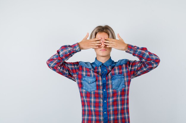 Portrait of young teen boy covering eyes with hands in checked shirt and looking scared front view