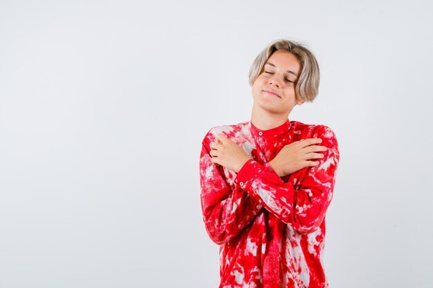 Portrait of young teen boy hugging himself, keeping eyes shut in shirt and looking delighted front view