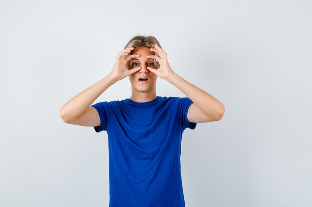 Portrait of young teen boy looking through fingers in blue t-shirt and looking astonished front view