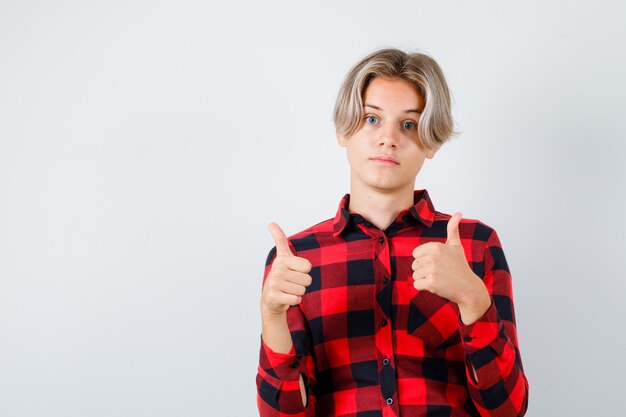 Портрет молодого мальчика-подростка показывает двойные пальцы вверх в клетчатой рубашке и выглядит довольным, вид спереди