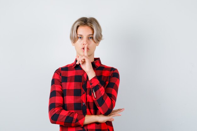 Portrait of young teen boy showing silence gesture in checked shirt and looking careful front view