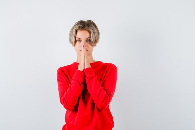 Portrait of young teen boy with hands in praying gesture in red sweater and looking hopeful front view